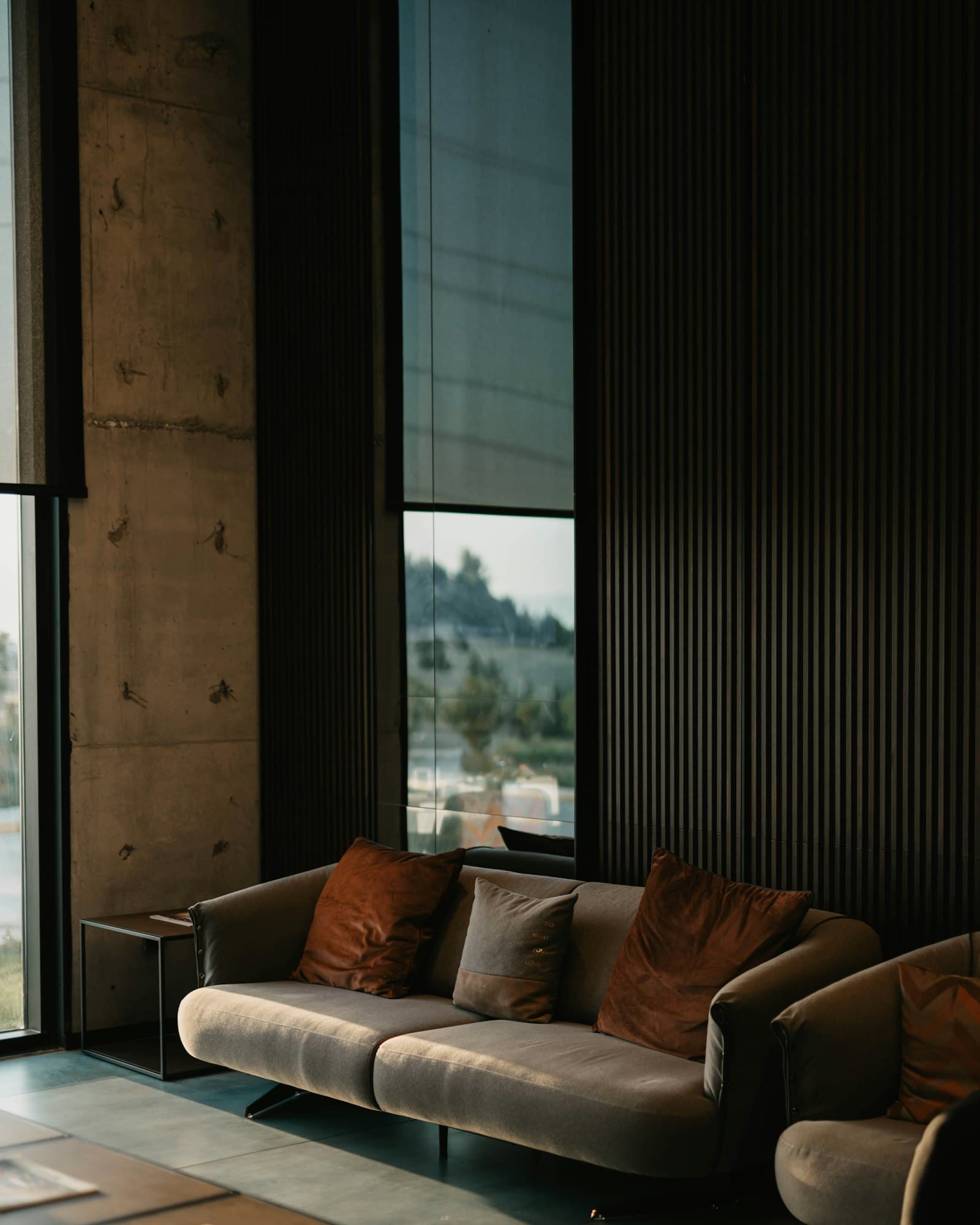 Loft interior in low warm light — board-formed concrete, full-height steel-framed glazing, and a wall of dark timber battens above a low sofa